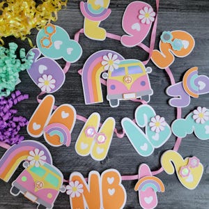 May include: A colorful banner with cut-out letters and shapes, including a rainbow, a flower-adorned van, and the letters "NB". The banner is decorated with flowers and hearts, and strung on a pink ribbon. The overall theme is a retro, hippie aesthetic.