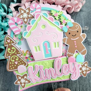 May include: A pink and green Christmas-themed decoration with a gingerbread house, gingerbread person, and the name "Kinley". The decoration includes candy canes, snowflakes, and a Christmas tree. The background has pink and blue ribbons.