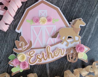 Puede incluir: Adorno para tarta en forma de granero rosa y marrón con un caballo, sombrero de vaquero y el nombre "Esther" en purpurina dorada. También se incluye el número "2". Decorado con flores de papel rosas y amarillas y hojas verdes.