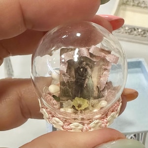 Peut inclure: Un globe en verre transparent avec une photographie vintage miniature à l'intérieur, orné de rubans roses et de petites pierres blanches. Le globe repose sur une base décorative rose et blanche, créant une esthétique nostalgique et délicate.