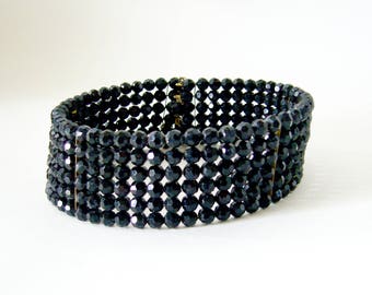 May include: A black choker necklace made of faceted glass beads. The beads are arranged in rows and are connected by gold-toned metal clasps.