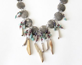May include: A silver necklace with a chain of round, textured discs and dangling charms. The charms include small, carved bone pieces, turquoise beads, and garnet beads.