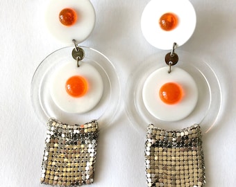 May include: A pair of white and orange statement earrings with a gold chainmail fringe. The earrings feature a white circle with an orange circle in the center, surrounded by a clear acrylic circle.