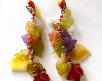 May include: A pair of long, dangle earrings featuring multiple layers of colorful plastic leaves in shades of green, yellow, orange, red, and purple. The leaves are attached to a gold-toned metal chain with small orange beads.