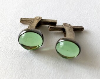 May include: A pair of silver cufflinks with green glass cabochons. The cufflinks are in the shape of a cross with a round cabochon in the center.