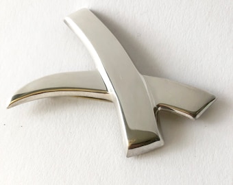 May include: A silver abstract brooch with a curved, angular design. The brooch is made of polished metal and has a smooth, reflective surface.