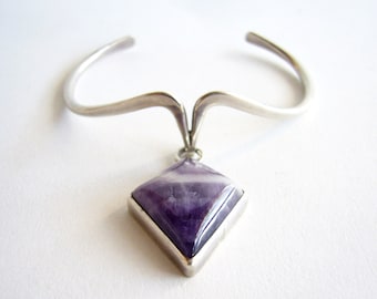 May include: A silver bangle bracelet with a dangling amethyst gemstone pendant. The pendant is a square shape with rounded corners and is set in a silver frame.