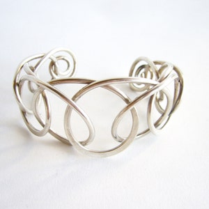 Puede incluir: Un brazalete plateado con un diseño abierto. El brazalete presenta una serie de bucles y espirales interconectados, creando un patrón intrincado y elegante. El metal tiene un acabado pulido, que refleja la luz y aumenta su atractivo visual.