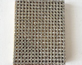 May include: A square-shaped compact with a gold frame and a surface covered in a grid pattern of small, clear rhinestones.