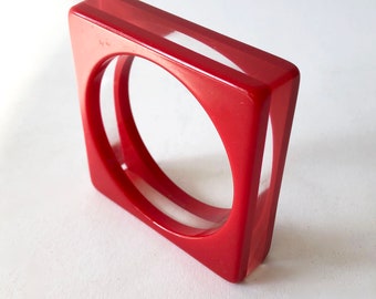 May include: A red square-shaped bangle with a clear plastic insert. The bangle has a double-layered design.