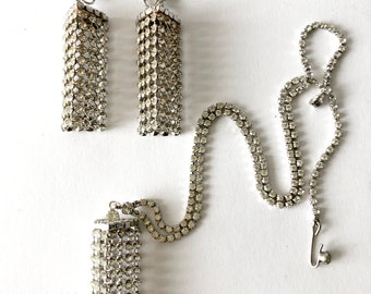 May include: A set of silver tone rhinestone jewelry featuring a necklace and a pair of clip-on earrings. The necklace and earrings have a dangling tassel design.