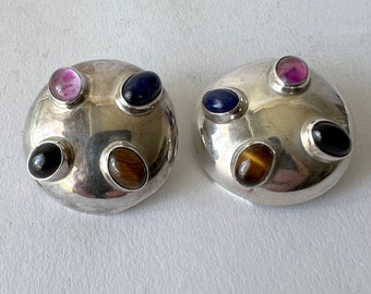 May include: A pair of silver earrings with five gemstones each. The gemstones are a mix of pink, blue, brown, and black.