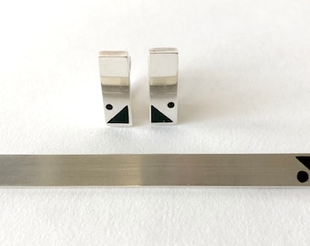 May include: A set of silver cufflinks and a tie bar with black geometric designs. The cufflinks are rectangular with a black triangle and a black circle on each. The tie bar is a long, thin rectangle with a black triangle and a black circle on one end.