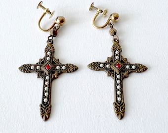 May include: A pair of gold tone clip-on earrings featuring ornate cross charms with white pearl accents and a red rhinestone in the center of each cross.