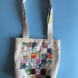 Puede incluir: Un bolso tote beige con un diseño de patchwork en la parte delantera. El patchwork presenta cuadrados de varias telas florales y estampadas en tonos de rojo, verde, azul y marrón. El bolso tiene dos asas con un estampado floral colorido.