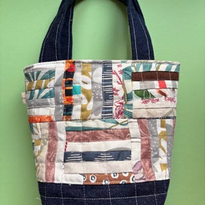 May include: A patchwork tote bag with a denim base and handles. The body of the bag is made of various fabric scraps in a variety of colours and patterns. The handles are a dark blue denim material.
