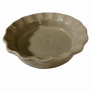 May include: A round, light beige ceramic dish with a smooth, glossy interior surface and a decorative, scalloped edge. The bowl features a shallow basin and a consistent, neutral-toned glaze that covers the entire visible surface of the piece.
