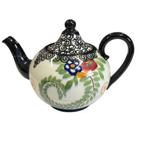 May include: A ceramic teapot with a cream-colored body, adorned with colorful floral designs and green foliage. The lid features a black, intricate pattern, and the spout and handle are also black. The teapot has a rounded shape and a glossy finish.