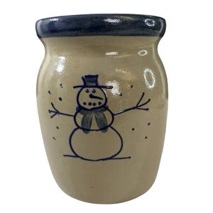 May include: A beige ceramic crock featuring a hand-painted dark blue snowman design on the front. The vessel has a wide, rounded body and a thick, dark blue glazed rim. The snowman illustration includes a top hat, scarf, and stick arms.