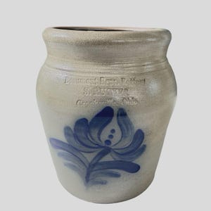 May include: A beige stoneware crock featuring a wide rim and a blue hand-painted floral design on the front. The surface displays stamped text reading "Burley and Winter Pottery", "BUTTER", and "Crooksville, Ohio" in a clear, indented font.