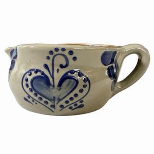 May include: A light grey ceramic creamer pitcher featuring a hand-painted blue heart design with decorative dots and swirls. The pitcher has a curved handle on the right side and a small pouring spout on the left, with a smooth, glazed finish.