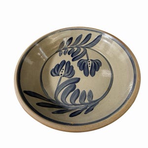 May include: A round ceramic bowl with a light beige base and a glossy finish. The interior features hand-painted dark blue floral motifs consisting of stylised leaves and two blossoms arranged within a circular border along the inner rim.