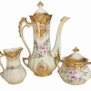 Pode incluir: Um conjunto de chá em porcelana composto por uma cafeteira alta com tampa, uma leiteira e um açucareiro com tampa. Cada peça apresenta uma base cor de creme com acabamentos dourados, bordas recortadas e um padrão floral de rosas.