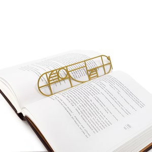 Gold Bauhaus-style metal bookmark lying flat across the pages of an open book — elegant and minimalist design for modern readers.