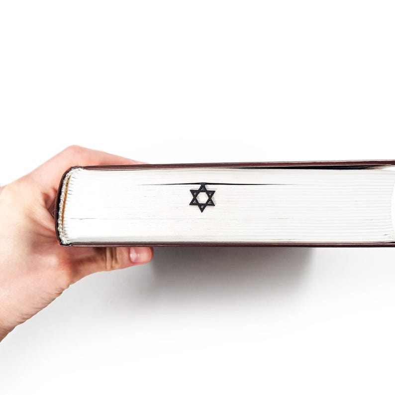 Close-up of a thick book with a black Star of David metal bookmark peeking out from the pages, held horizontally by a hand, clean white background.