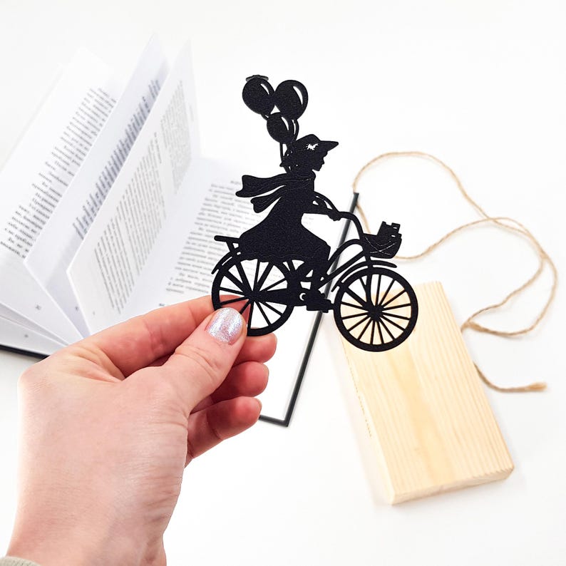 Hand holding a black metal bike bookmark shaped as a girl riding a bicycle with balloons, with an open book and a wooden engraved block in the background on a white surface.