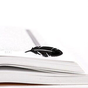 May include: A black feather bookmark on top of a white book with pages turned down.