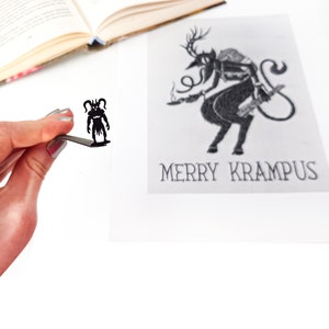 Puede incluir: Una ilustración en blanco y negro de Krampus, una criatura mítica con cuernos, cola y un saco en la espalda. La ilustración está en una tarjeta blanca con el texto "Merry Krampus".