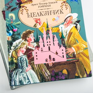 A pink metal castle bookmark with a book in the background. The book cover is blue and has a picture of a man and two children.