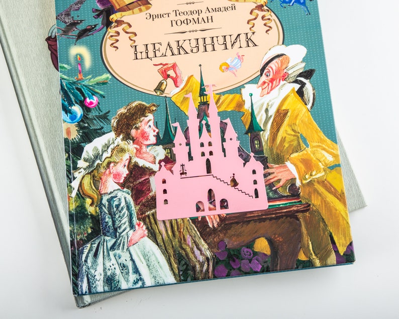 A pink metal castle bookmark with a book in the background. The book cover is blue and has a picture of a man and two children.