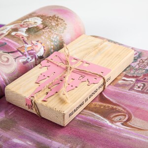 A wooden packaging block with a  with a pink metal castle cutout bookmark and a twine bow. The block has the text 'Reading is good for you' printed on it.