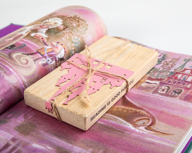A wooden packaging block with a  with a pink metal castle cutout bookmark and a twine bow. The block has the text 'Reading is good for you' printed on it.