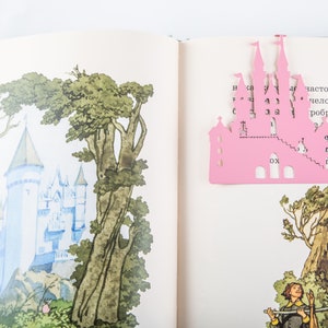 Pink metal bookmark Princess Castle. The large pink castle is between the pages of the book. The little crown is on the side of the book. It is visible even when the book is closed. Made in Ukraine. Not mass manufactured.