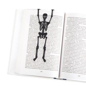 Skeleton bookmark. Premium quality book accessory laser cut from high-grade steel, powder coated by black polymer paint. The large meat skeleton stays between the pages of the book. The tips of his fingers are visible on the side of the book.