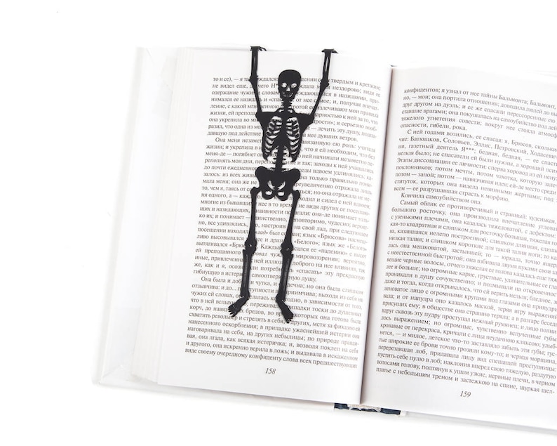 Skeleton bookmark. Premium quality book accessory laser cut from high-grade steel, powder coated by black polymer paint. The large meat skeleton stays between the pages of the book. The tips of his fingers are visible on the side of the book.