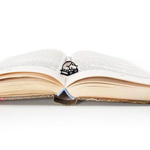 Op de afbeelding: Zwart metalen boekenlegger met een slapende kat op boeken, geplaatst in een open boek. Het boek heeft vergeelde pagina's en een zichtbare rug. Een blauw lint markeert de pagina. Een charmant accessoire.