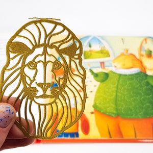 May include: A gold-coloured metal bookmark shaped like a lion's head with a detailed outline of the mane and face.
