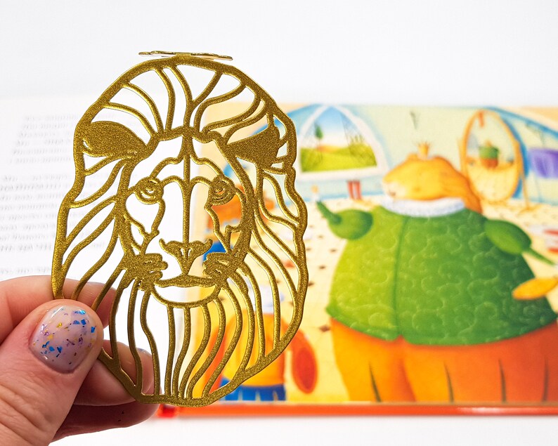 May include: A gold-coloured metal bookmark shaped like a lion's head with a detailed outline of the mane and face.
