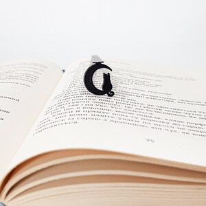 May include: Black metal bookmark shaped like a cat sitting on a crescent moon. The bookmark is a decorative accessory for book lovers.