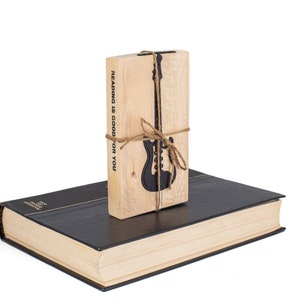 Electric guitar-shaped black metal bookmark tied to a rustic wooden block stamped with Reading is good for you, placed on a closed book. A fun gift for rock-loving readers.