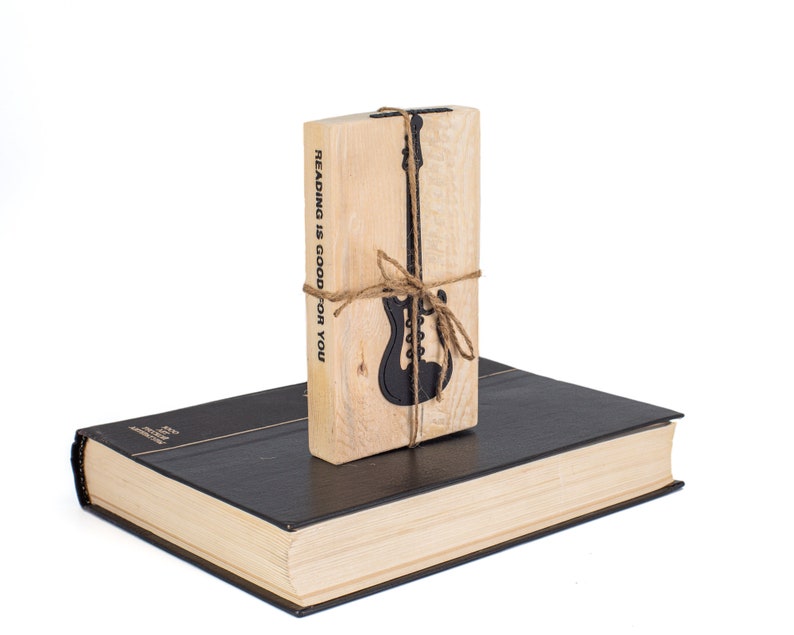 Electric guitar-shaped black metal bookmark tied to a rustic wooden block stamped with Reading is good for you, placed on a closed book. A fun gift for rock-loving readers.