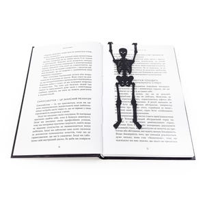 A black metal skeleton-shaped bookmark lying across the center of an open book, posed as if climbing up the pages.