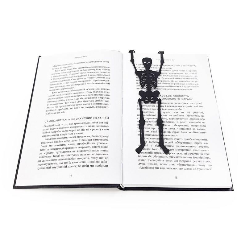 A black metal skeleton-shaped bookmark lying across the center of an open book, posed as if climbing up the pages.