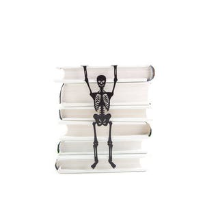 A black metal skeleton-shaped bookmark hanging between the pages of a stacked pile of white books, as if climbing up the stack.