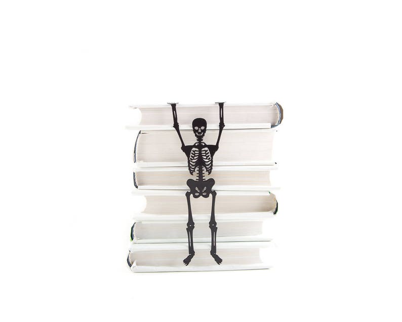 A black metal skeleton-shaped bookmark hanging between the pages of a stacked pile of white books, as if climbing up the stack.
