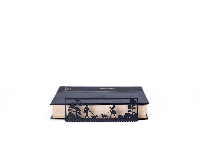 May include: Black metal bookmark with a silhouette design of two people walking dogs in a park. The bookmark is placed on a closed book.